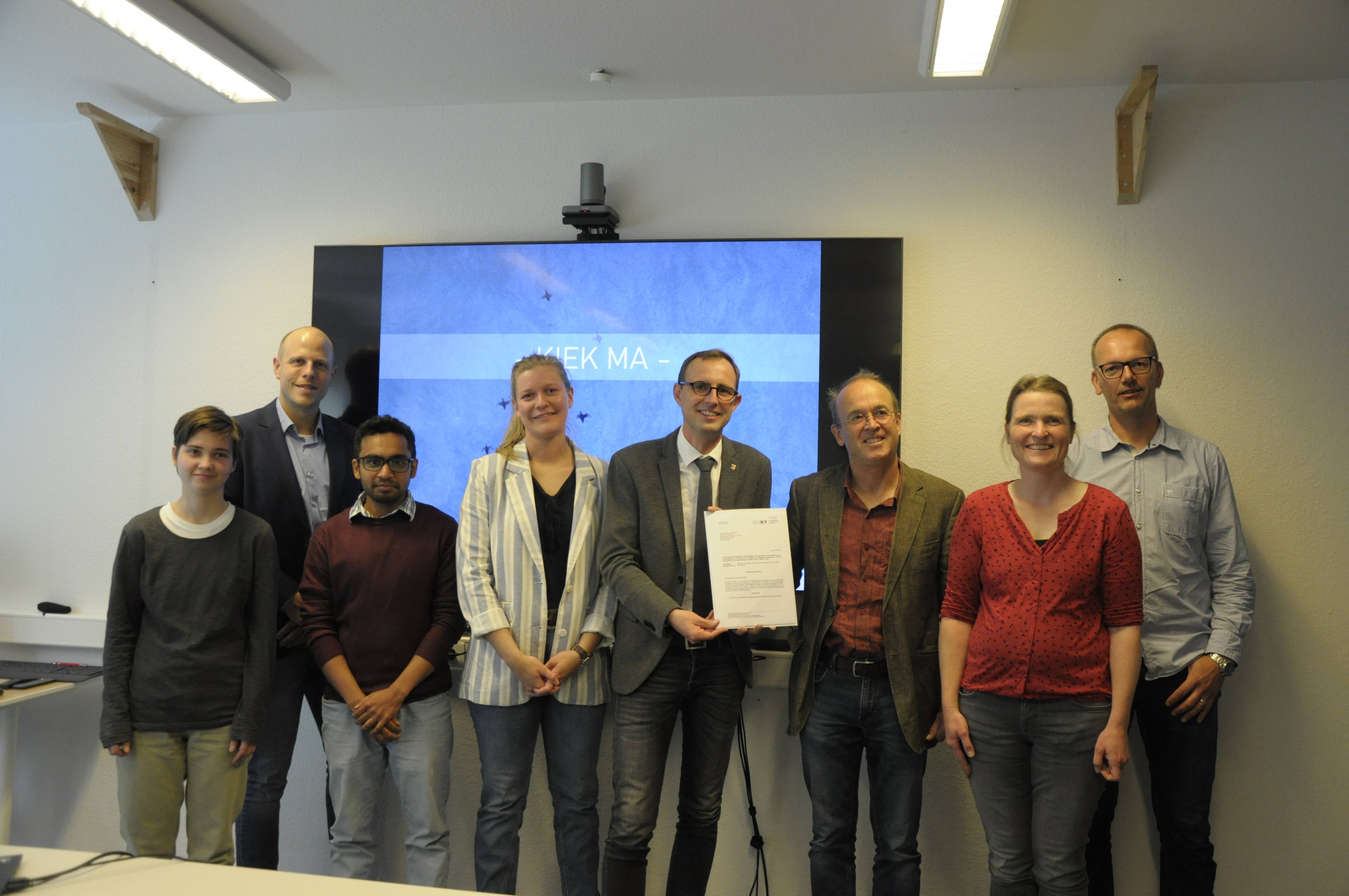Übergabe des Zuwendungsbescheid durch Dirk Schrödter. Die Gruppe von Eco Consult steht vor einem Monitor. In der Mitte der Minister mit dem Bescheid.